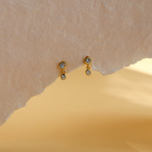 Pair of tiny gold bezel-set CZ drop studs, Leda earrings by Rainbow Rocks, on textured paper